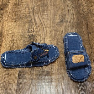 以下が含まれることがあります： フリンジの付いた青いデニムのスリッパのペア。スリッパには、片方のスリッパに「Levi's Jeans」という文字が書かれた茶色の革のパッチ、もう片方のスリッパにポケットが付いています。