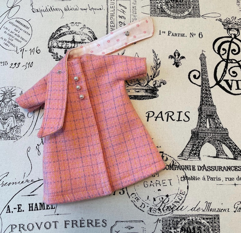Puede incluir: Un conjunto de vestido y chaqueta de mu&ntilde;eca a cuadros rosas con botones perlados. La chaqueta tiene un cuello a juego con detalles de estrellas. El conjunto est&aacute; dise&ntilde;ado con un estilo cl&aacute;sico, perfecto para el guardarropa de una mu&ntilde;eca.