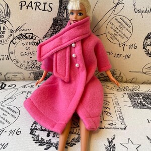 May include: A pink doll wearing a pink coat with a matching scarf and pearl buttons. The doll has blonde hair and blue eyes. The doll is sitting on a white fabric background with a Parisian theme.