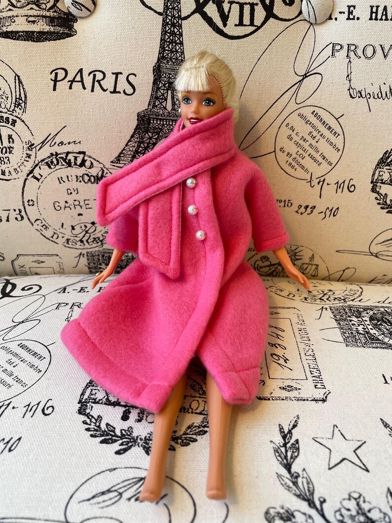 May include: A pink doll wearing a pink coat with a matching scarf and pearl buttons. The doll has blonde hair and blue eyes. The doll is sitting on a white fabric background with a Parisian theme.