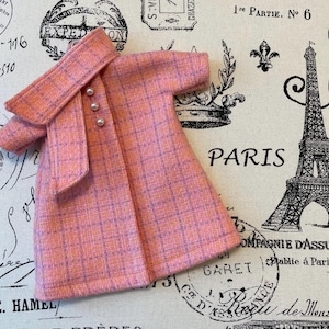 Puede incluir: Un abrigo rosa a cuadros con cuello a juego y tres botones de perlas. El abrigo es de color rosa claro con un sutil patr&oacute;n de cuadr&iacute;cula p&uacute;rpura. El abrigo se muestra sobre una tela con la palabra "PARIS" y una Torre Eiffel.