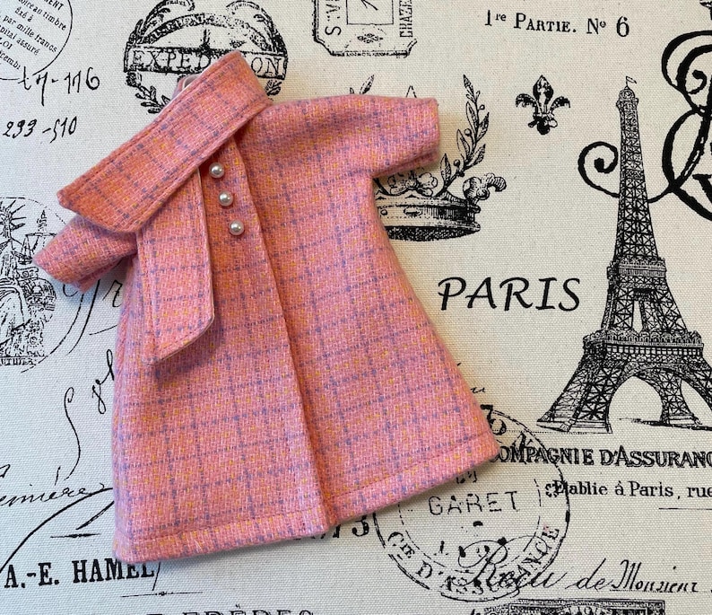 Puede incluir: Un abrigo rosa a cuadros con cuello a juego y tres botones de perlas. El abrigo es de color rosa claro con un sutil patr&oacute;n de cuadr&iacute;cula p&uacute;rpura. El abrigo se muestra sobre una tela con la palabra "PARIS" y una Torre Eiffel.