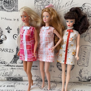 May include: Three fashion dolls standing side-by-side. The left doll has blonde hair and wears a pink and red geometric print dress. The center doll has light brown hair and wears a pink and white grid print dress. The right doll has dark brown hair and wears a white dress with a yellow floral pattern and an orange vertical stripe.
