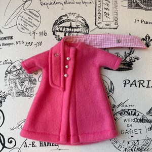 May include: A pink fleece coat with a white and pink gingham scarf. The coat has a button closure and pearl-like buttons.