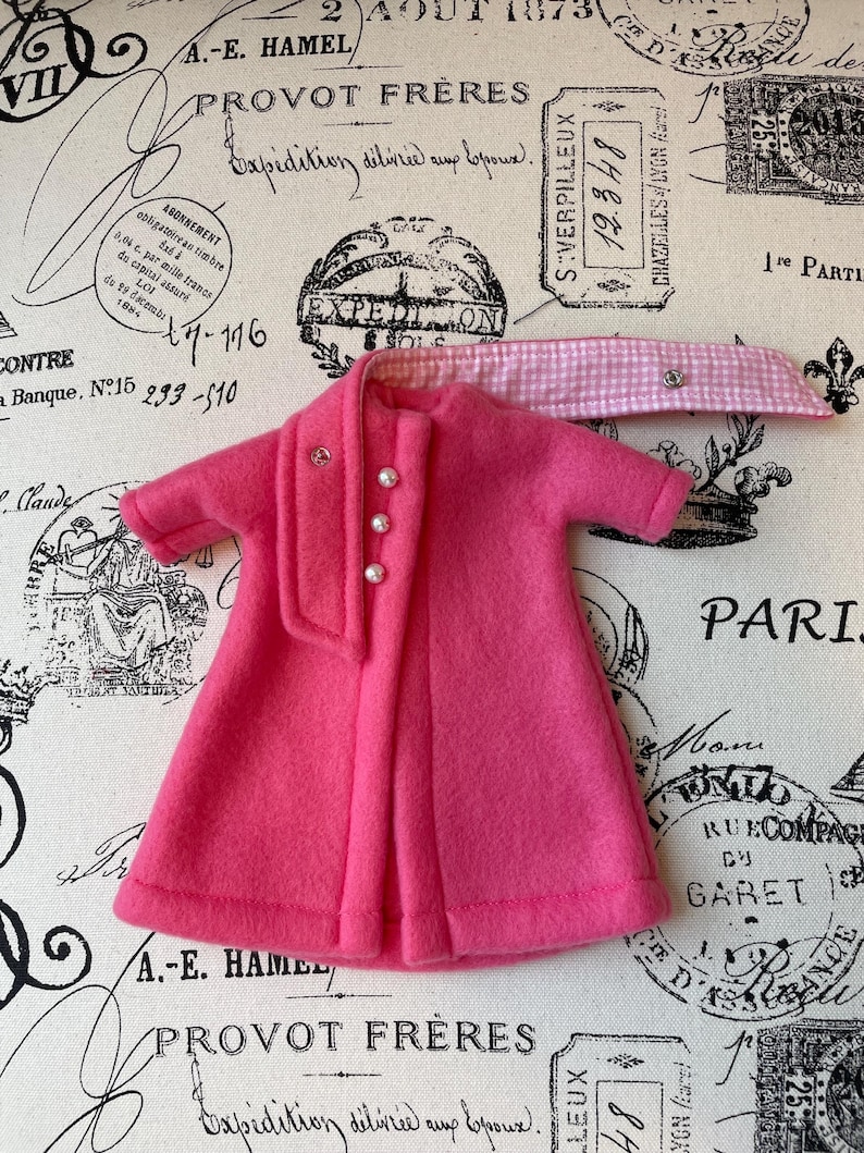 May include: A pink fleece coat with a white and pink gingham scarf. The coat has a button closure and pearl-like buttons.