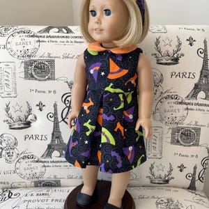 Könnte beinhalten: Eine Puppe mit blondem Haar trägt ein schwarzes Stirnband und ein ärmelloses Kleid mit einem Muster aus orangefarbenen, violetten und grünen Hexenhüten und Fledermäusen auf schwarzem Grund, dazu ein einfarbiger orangefarbener Kragen und schwarze flache Schuhe.