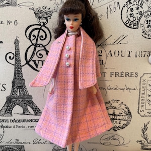 Puede incluir: Una mu&ntilde;eca vintage con un abrigo a cuadros rosa y naranja con un chal a juego. El abrigo tiene tres botones de perla y una peque&ntilde;a broche. La mu&ntilde;eca tiene cabello casta&ntilde;o y lleva un collar de perlas. El fondo presenta escritura y sellos franceses.