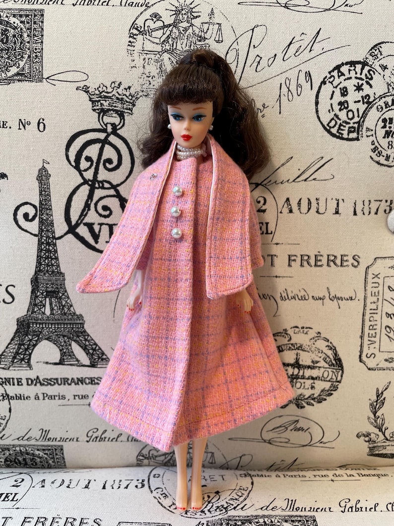 Puede incluir: Una mu&ntilde;eca vintage con un abrigo a cuadros rosa y naranja con un chal a juego. El abrigo tiene tres botones de perla y una peque&ntilde;a broche. La mu&ntilde;eca tiene cabello casta&ntilde;o y lleva un collar de perlas. El fondo presenta escritura y sellos franceses.
