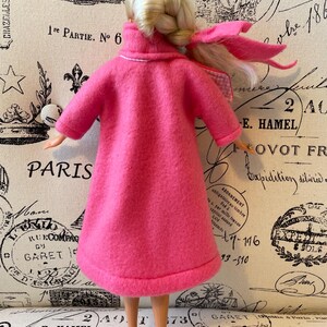 May include: A pink fleece coat with a white trim around the collar and cuffs. The coat is worn by a doll with blonde hair and a pink ribbon.