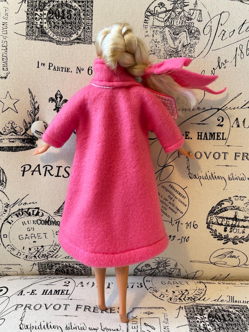 May include: A pink fleece coat with a white trim around the collar and cuffs. The coat is worn by a doll with blonde hair and a pink ribbon.