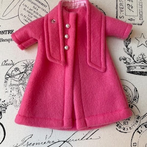 May include: A pink fleece coat with a white and pink checkered lining and pearl buttons. The coat has a shawl collar and long sleeves.