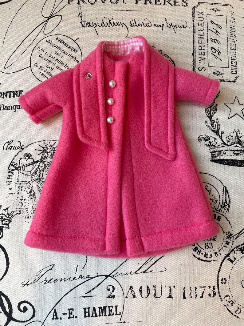 May include: A pink fleece coat with a white and pink checkered lining and pearl buttons. The coat has a shawl collar and long sleeves.