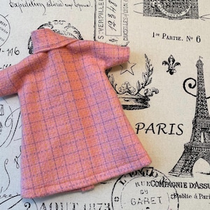 Puede incluir: Un abrigo de mu&ntilde;eca rosa y morado a cuadros con cuello y mangas cortas. El abrigo est&aacute; hecho de una tela texturizada. El fondo presenta una tela con tem&aacute;tica parisina con la Torre Eiffel y texto.