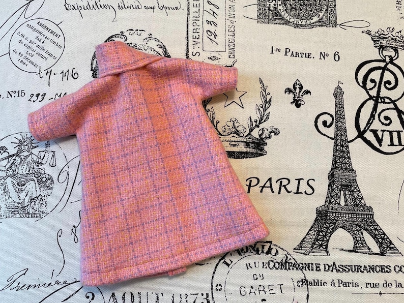 Puede incluir: Un abrigo de mu&ntilde;eca rosa y morado a cuadros con cuello y mangas cortas. El abrigo est&aacute; hecho de una tela texturizada. El fondo presenta una tela con tem&aacute;tica parisina con la Torre Eiffel y texto.