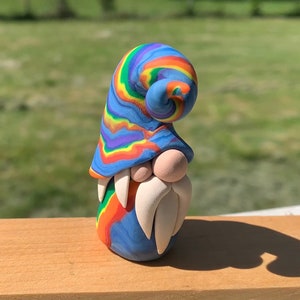 May include: A colourful gnome figurine made from clay. The gnome has a blue hat with rainbow swirls, a beige nose, and a long white beard. The body is a mix of blue, orange, and green. This decorative piece is ideal for adding a touch of fantasy to a home.