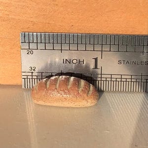 Könnte beinhalten: Ein Miniatur-Brotlaib, etwa 2,5 cm lang, mit hellbrauner Kruste und detaillierter Riffelung. Ein Edelstahl-Lineal liegt zur Größenbestimmung neben dem Brot, was auf ein Puppenhaus- oder Miniatur-Lebensmittel-Objekt hindeutet.