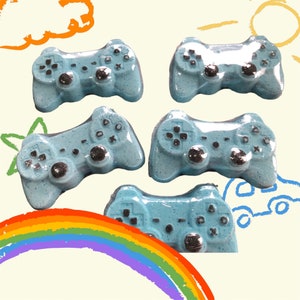 May include: Four blue bath bombs shaped like video game controllers. The bath bombs have silver accents and are on a white background.