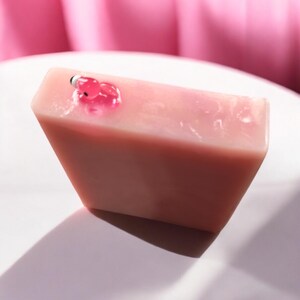 May include: A pink, rectangular soap bar with a small, translucent pink and black decorative element on top. The soap has a smooth surface. The soap is set against a white background with a pink backdrop.