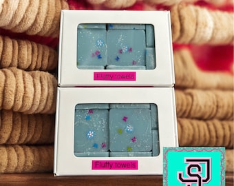 Könnte beinhalten: Zwei weiße Boxen mit klaren Fenstern, die blaue Wachsschmelzen mit weißen Blüten und buntem Glitzer zeigen. Die Boxen sind übereinander gestapelt und haben den Text "Fluffy towels" aufgedruckt. Ein türkisfarbenes Logo mit dem Text "S.J's Snazzy Soaps & candle co." ist in der unteren rechten Ecke sichtbar.