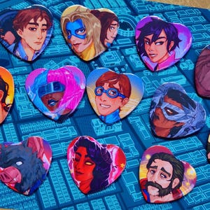 May include: A collection of heart-shaped pins featuring various illustrated characters with diverse facial features, hairstyles, and colorful outfits, arranged on a blue surface with a grid pattern of city streets and buildings.