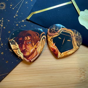 Puede incluir: Dos pines en forma de corazón con ilustraciones detalladas. Uno presenta a un hombre con cabello castaño y barba, el otro una figura encapuchada. Ambos tienen detalles dorados y anaranjados. Descansan sobre una superficie de madera con fondo azul marino.