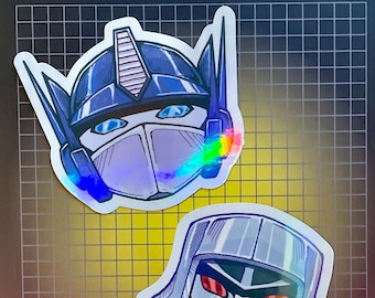 May include: Two holographic stickers featuring the heads of Optimus Prime and Megatron from Transformers. Optimus Prime is blue and white, and Megatron is silver and gray. The background has the Transformers logo and the phrase "More Than Meets The Eye!"