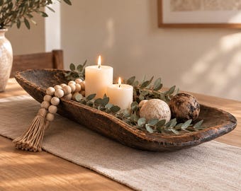May include: A long, dark brown, hand-carved wooden dough bowl containing two lit white pillar candles, a strand of light-colored wooden beads with a tassel, green eucalyptus sprigs, and two textured decorative spheres in beige and dark brown.