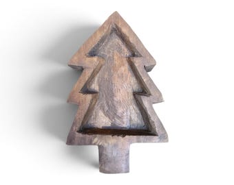 May include: A brown wooden bowl carved into the shape of a pine tree. The interior of the bowl features a recessed, tiered silhouette that mirrors the outer triangular form of the tree, with a flat base and a central trunk-like extension at the bottom.