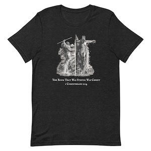 Può includere: T-shirt grigio scuro con un'illustrazione in bianco e nero di una scena religiosa, con una figura sulla croce. Il testo "The Rock That Was Struck Was Christ 1 Corinzi 10:4" è stampato sotto.