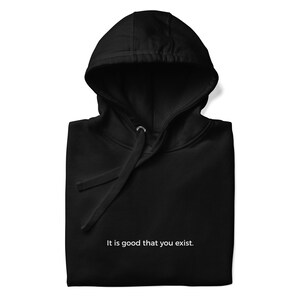 May include: Black hooded sweatshirt with a drawstring and the text "It is good that you exist." The sweatshirt is folded and has a simple, minimalist design. The fabric appears soft and comfortable.