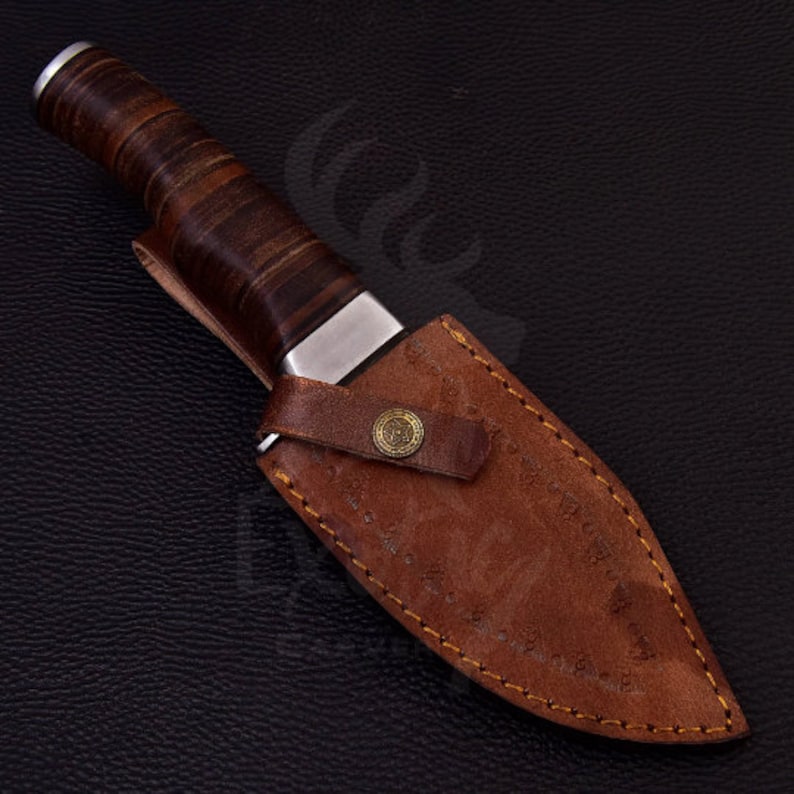 Puede incluir: Una funda de cuero marr&oacute;n con un borde cosido y un bot&oacute;n de metal. Un mango de cuchillo envuelto en cuero marr&oacute;n es visible dentro de la funda.