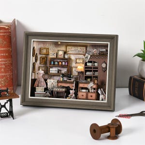 Könnte beinhalten: Gerahmtes Miniatur-Diorama eines Nähladens. Die Szene umfasst eine Schneiderpuppe mit einem rosa Kleid, Nähmaschinen, Regale mit Garnrollen und einen kleinen Teddybären. Der Rahmen ist grau.