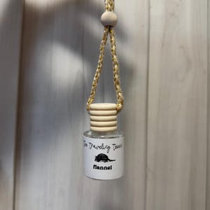 May include: A small, clear glass bottle with a wooden cap and a braided cord for hanging. The bottle has a label with the text "The Traveling Texas" and "flannel" with a cartoon illustration of a rodent.