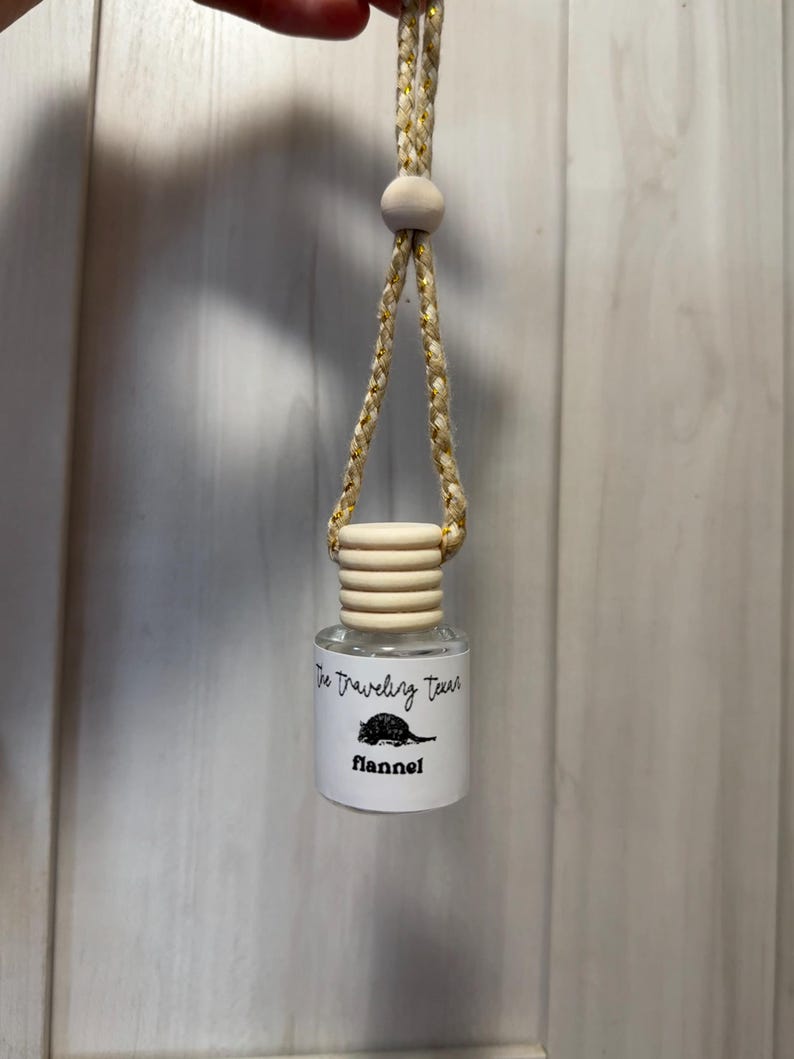 May include: A small, clear glass bottle with a wooden cap and a braided cord for hanging. The bottle has a label with the text "The Traveling Texas" and "flannel" with a cartoon illustration of a rodent.