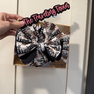 Peut inclure: Un nœud pour bébé fait à la main avec un motif unique noir, blanc et marron. Le nœud est fixé sur une planche de bois avec le texte "The Traveling Texan" en rose et noir.