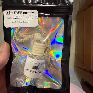May include: An air freshener in a black bag with a holographic interior. The label reads "Air Diffuser" and "Cowgirl britches" scent. The bottle has a wooden top and a string for hanging. The text "The Traveling Texan" is also visible.