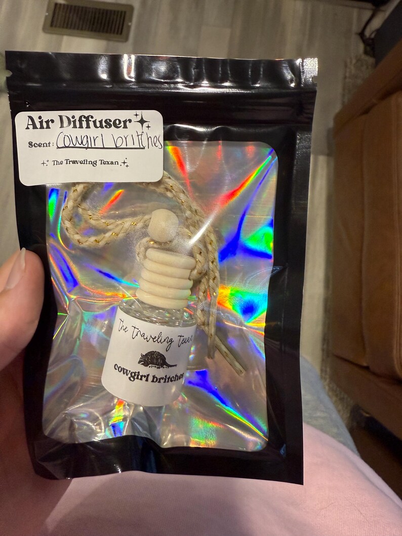May include: An air freshener in a black bag with a holographic interior. The label reads "Air Diffuser" and "Cowgirl britches" scent. The bottle has a wooden top and a string for hanging. The text "The Traveling Texan" is also visible.