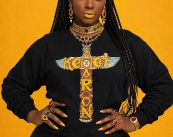 May include: A woman wearing a black sweatshirt with the word "Kemet Aaron" printed on it. She is wearing a gold crown, earrings, and necklaces. Her nails are painted yellow, and she is wearing several rings. The background is yellow.
