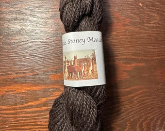 Puede incluir: Ovillo de hilo marrón oscuro con una etiqueta que dice "Stoney Meadows". La etiqueta presenta una escena pastoral con llamas. El hilo está bien enrollado y parece suave, ideal para proyectos de tejido o ganchillo. El hilo se mide probablemente en gramos.