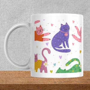 Op de afbeelding: Witte keramische mok met een kleurrijk kattenpatroon. Het ontwerp bevat verschillende kattenillustraties in oranje, paars, geel en groen, met hartjes en bloemmotieven. De mok heeft een gebogen handvat.