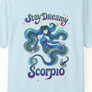 以下が含まれることがあります： 水色のTシャツ。長い髪の女性、サソリ、そして「Stay Dreamy Scorpio」の文字が描かれたカラフルなイラストが特徴です。デザインには、青、緑、紫、ピンクの色合いが使用されています。