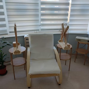 May include: A light wood chair with a white cushion is centered in a room with two small wooden side tables. Each table holds a wooden structure. A potted plant is on the left, and a device on a small table is on the right.