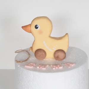 May include: A yellow toy duck with brown wheels and an orange beak. The duck has a string attached and is sitting on a white surface. Pink letters spell out the word "BABY".