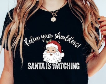 May include: Black t-shirt with the text "Relax your shoulders! SANTA IS WATCHING" and a graphic of Santa Claus. The Santa graphic features a red hat, white beard, and a smiling face. The shirt is paired with dark gray jeans and white sneakers.