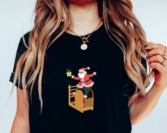 May include: Black t-shirt with a festive Santa Claus graphic. The design depicts Santa on a wooden structure, holding a present. The colours used are red, white, and green. The shirt has a classic crew neck.