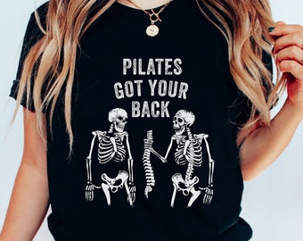 以下が含まれることがあります： 「PILATES GOT YOUR BACK」の文字と、背骨を持つ2体の骸骨のグラフィックがプリントされた黒いTシャツ。画像の下部には「BLACK」の文字がプリントされています。Tシャツは人が着ています。