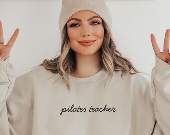 May include: A beige sweatshirt with the words "pilates teacher" in black script. The sweatshirt is paired with a matching beanie. The person is making a peace sign with their fingers.