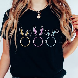 Könnte beinhalten: Schwarzes T-Shirt mit einem Motiv von drei Ostereiern mit Hasenohren. Die Eier sind in Gelb, Rosa und Blau umrandet.