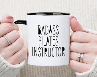 May include: White ceramic mug with a black rim and handle. The mug has black text that reads "BADASS PILATES INSTRUCTOR".
