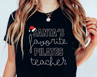 May include: Black t-shirt with the text "Santa's favorite Pilates teacher" in white script. A small Santa hat graphic is above the word "favorite". The shirt is paired with dark wash jeans and white sneakers. A festive holiday themed shirt.